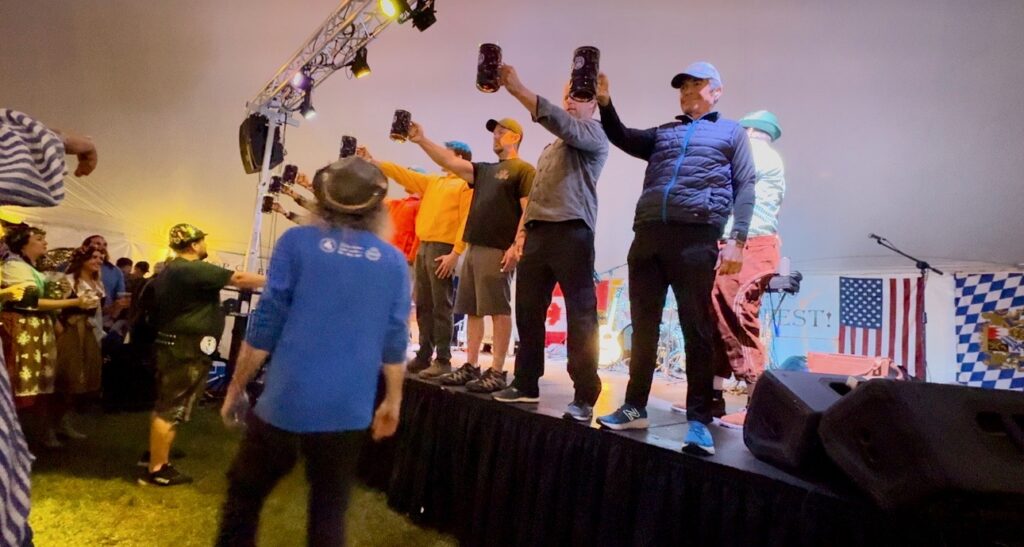 Seven men standing on a stage holding beer steins